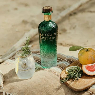 eine grüne Flasche Mermaid Gin steht auf einem Tusch am Strand. 